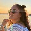 Woman with curly hair wearing sunglasses and smiling at sunset by the water.