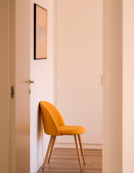 Orange upholstered chair with wooden legs against a white wall under framed artwork.