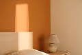 Bedside table with a lamp and sunlight casting shadows on a plain beige wall.