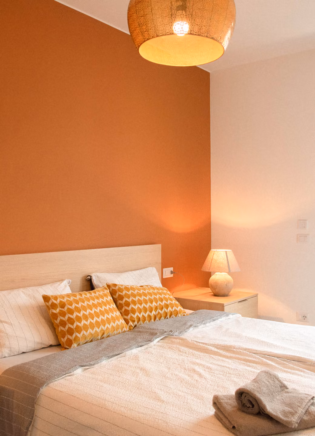 Modern bedroom with white bed, patterned orange pillows, bedside lamp, and warm orange accent wall.