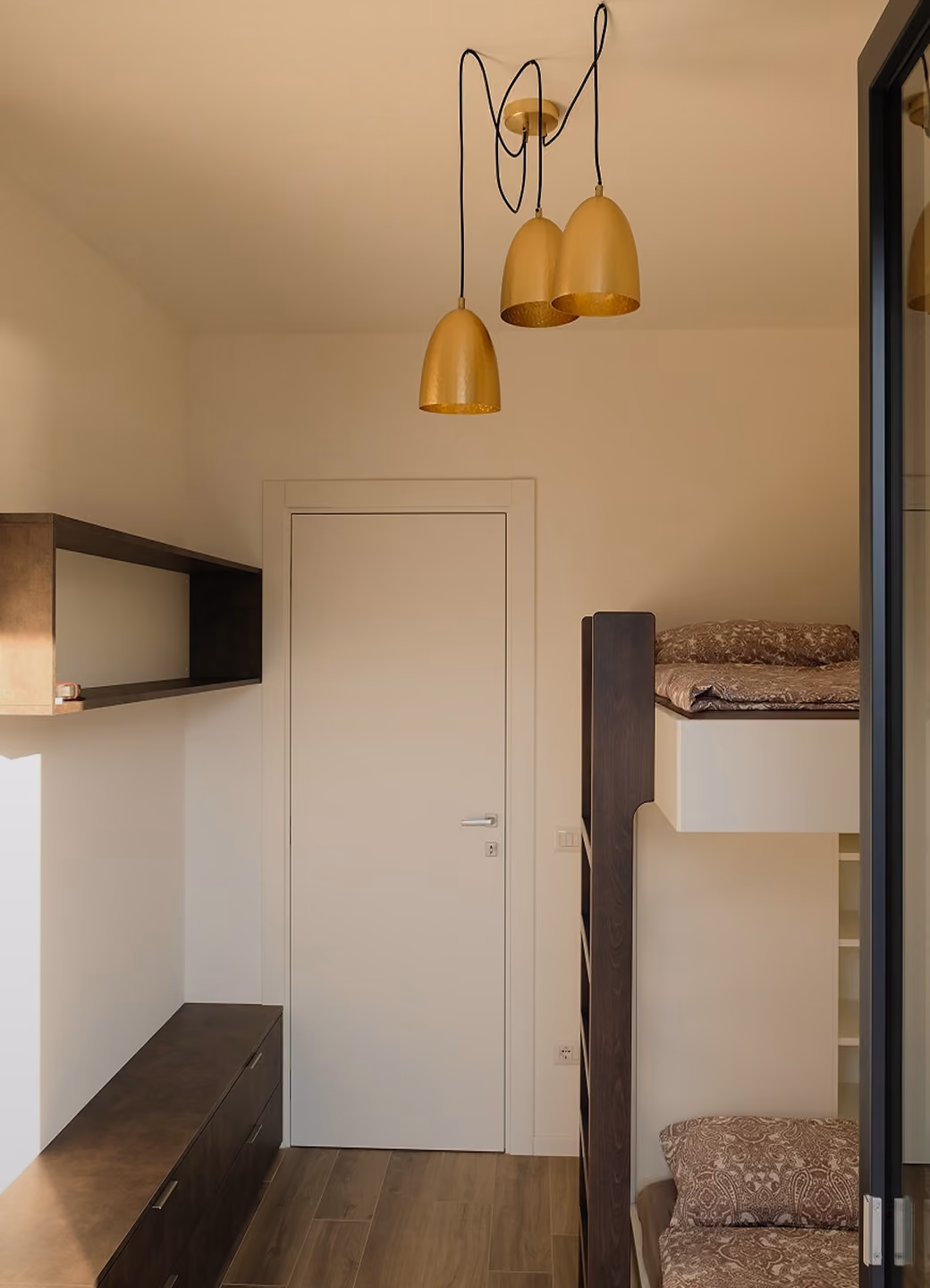 Small room with wooden bunk bed, patterned bedding, dark wooden shelves, and three gold hanging lamps.