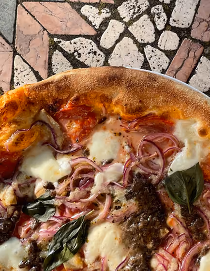 Close-up of a pizza with melted cheese, red onions, basil leaves, and dark sauce on a stone table.