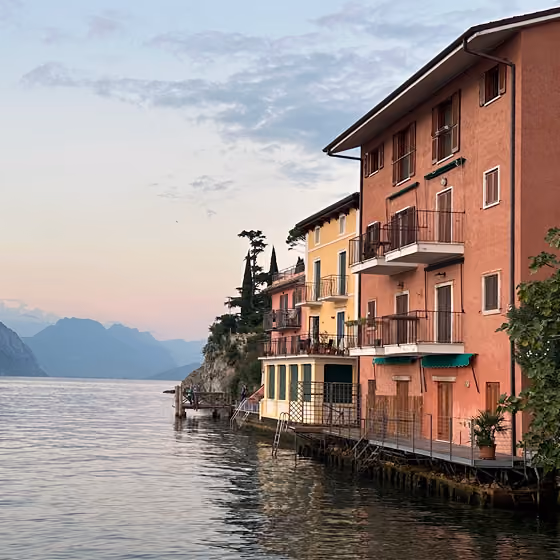 Colorful lakeside buildings with balconies and mountain views at sunset under a partly cloudy sky.