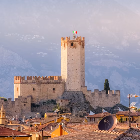 Historic stone castle with a tall tower flying the Italian flag above rooftops and mountains in the background.