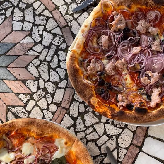 Two pizzas on a patterned stone table; one with sausage and red onions, the other with cheese and toppings.