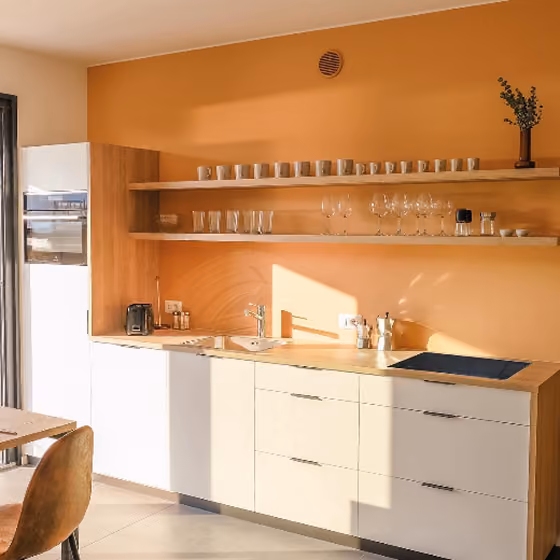 Modern kitchen with orange walls, wooden shelves holding glasses and mugs, and white cabinets.