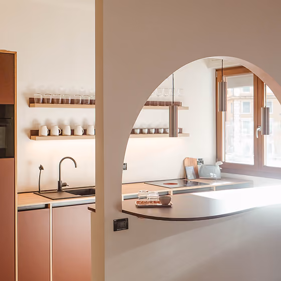 Modern kitchen with an arched pass-through, wooden shelves holding mugs and glasses, and large windows.
