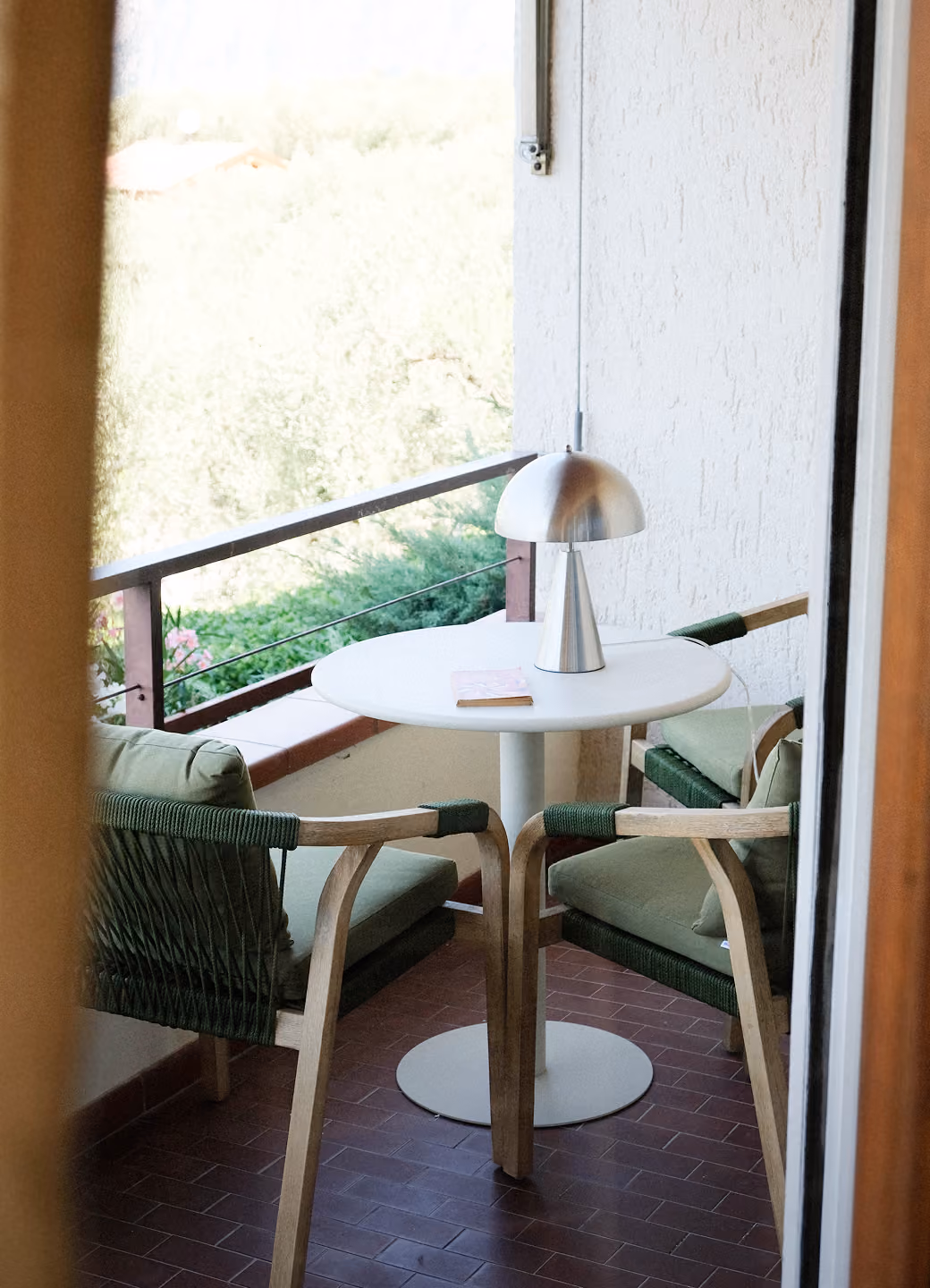 Balcony with two green cushioned chairs around a small white round table with a silver lamp and book.