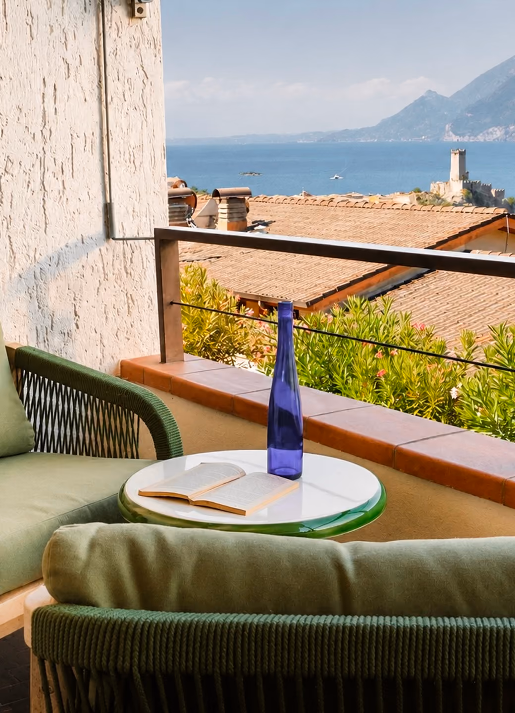 Balcony with green cushioned chairs, small round table, blue bottle, open book, and sea view.