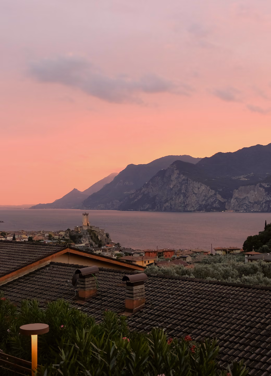 Sunset over a lakeside town with tiled roofs, a tower, and mountains in the background.