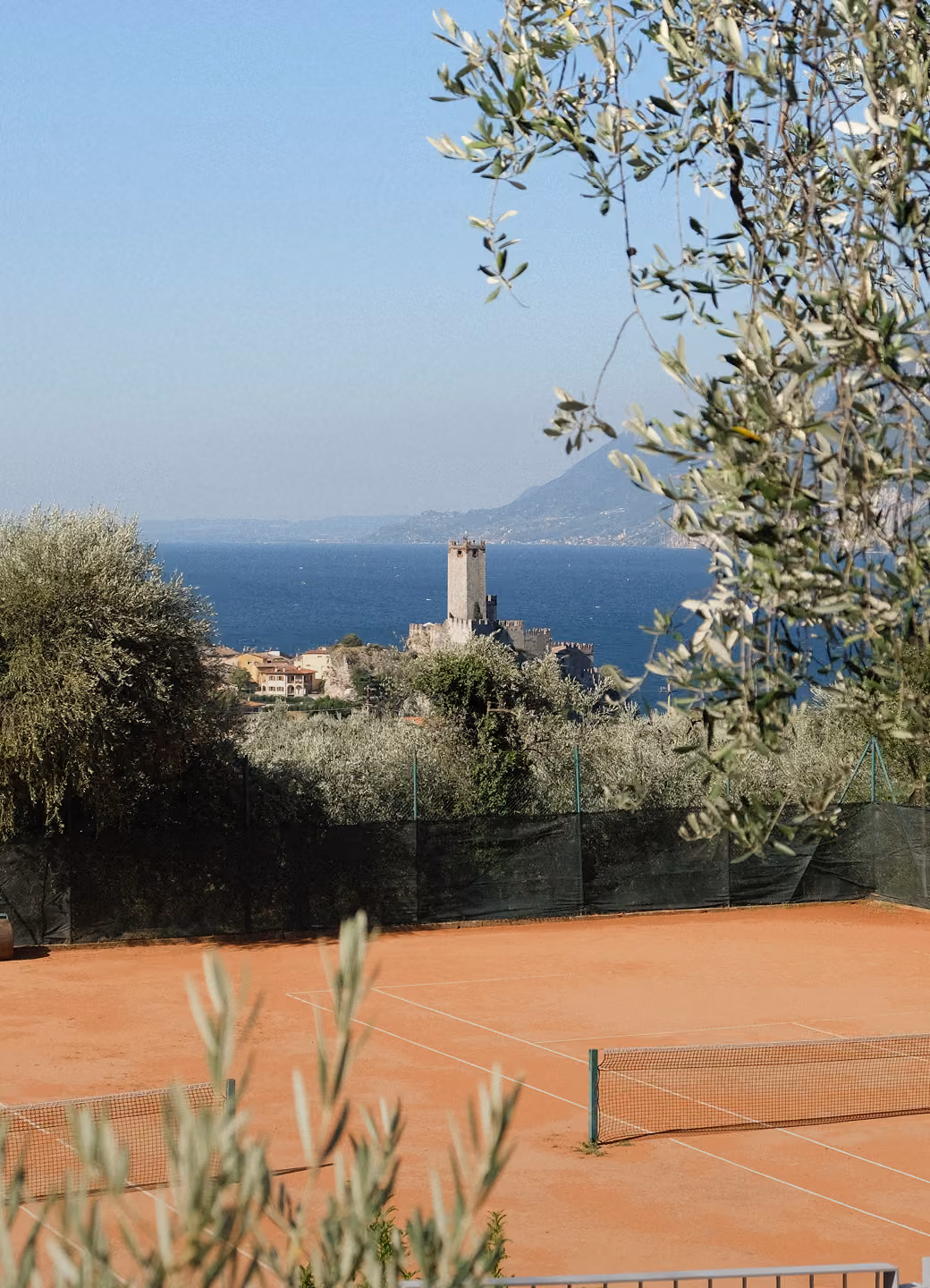 Clay tennis court with net in foreground, olive trees, a castle, and a blue lake with mountains behind.