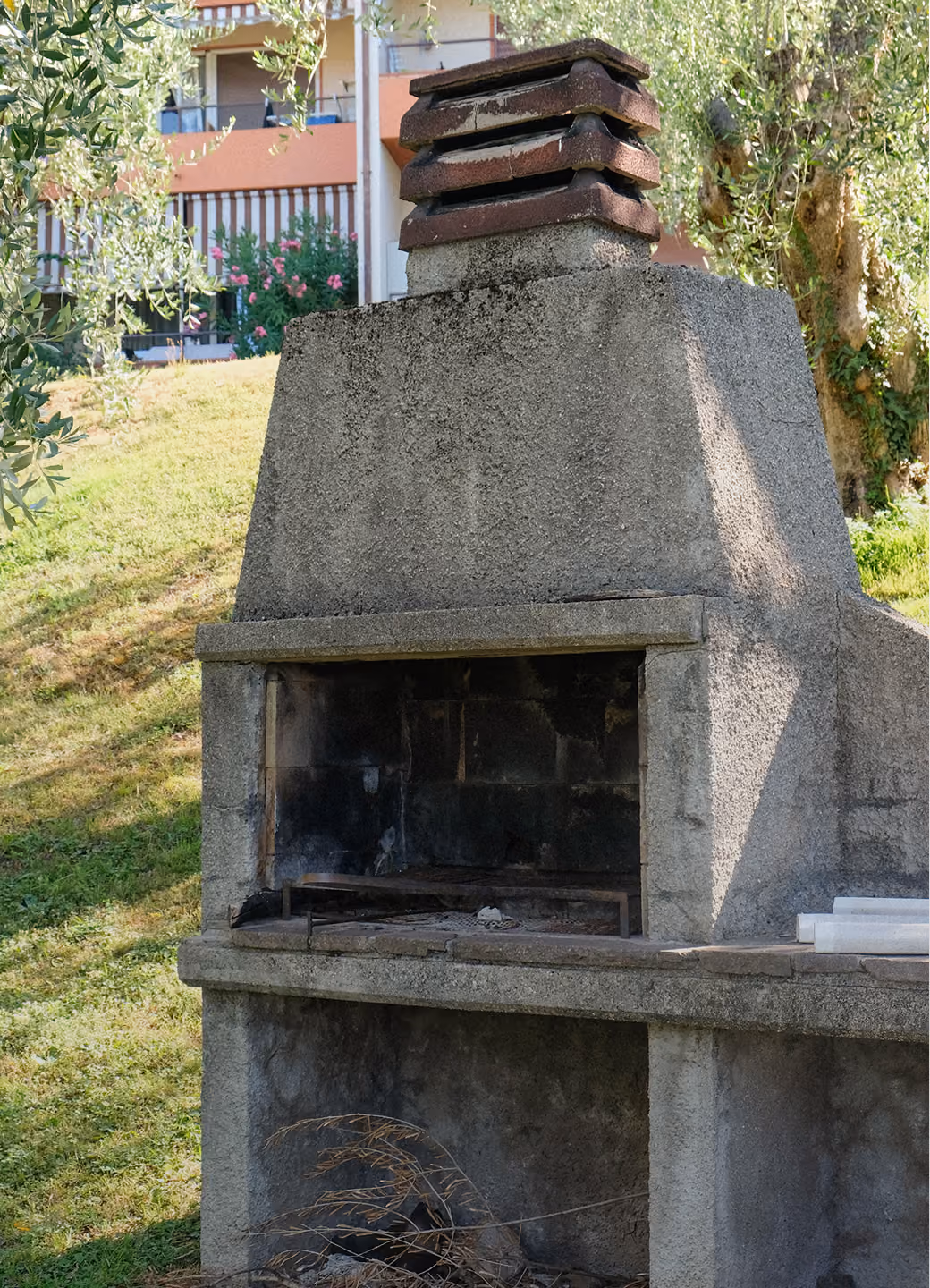 Outdoor concrete barbecue grill with chimney in a sunny garden setting near a building.