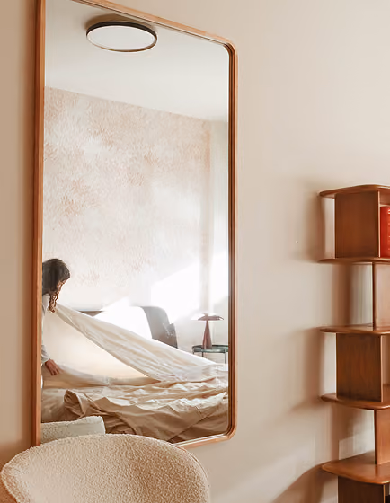Wood-framed mirror reflects a woman making a bed in a softly lit, cozy bedroom.