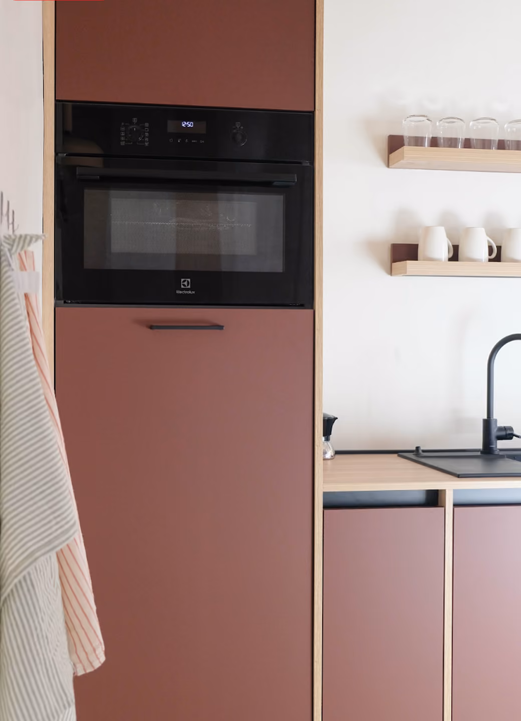 Built-in black oven mounted in brown kitchen cabinetry with black faucet and shelves holding glassware and pitchers.