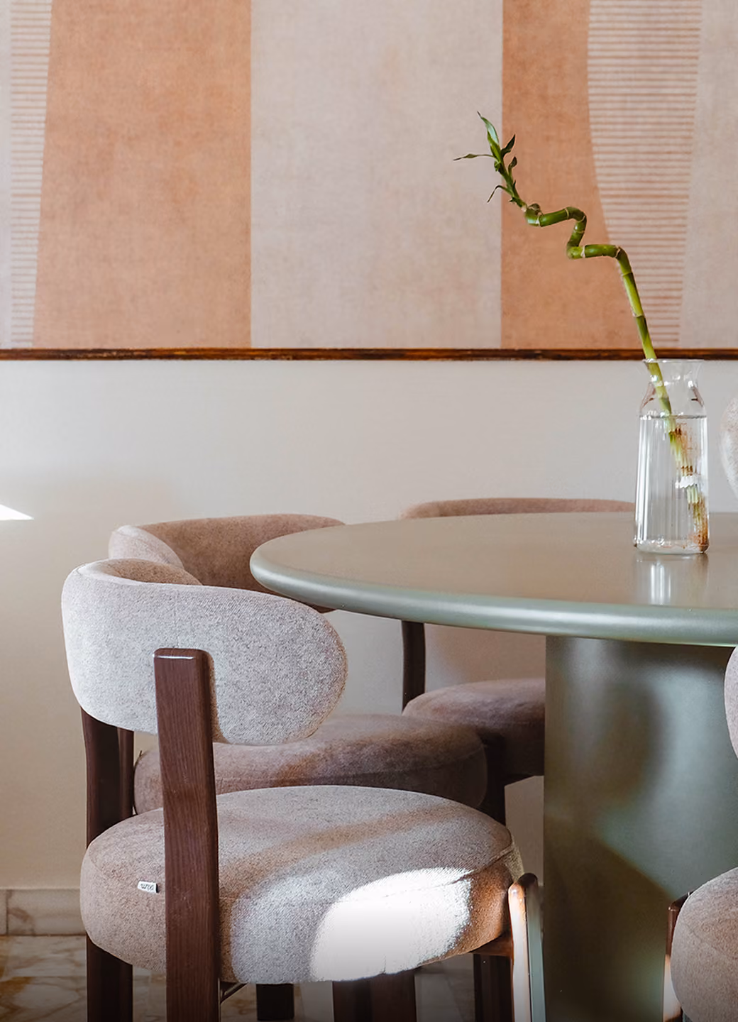 Round table with padded chairs and a glass vase holding a curved bamboo stalk in a modern room.