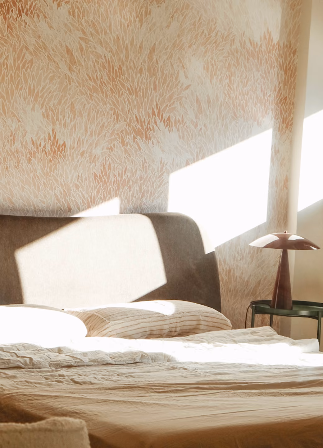 Sunlight casts shadows on a beige bed with striped pillows and a bedside table with a mushroom-shaped lamp.