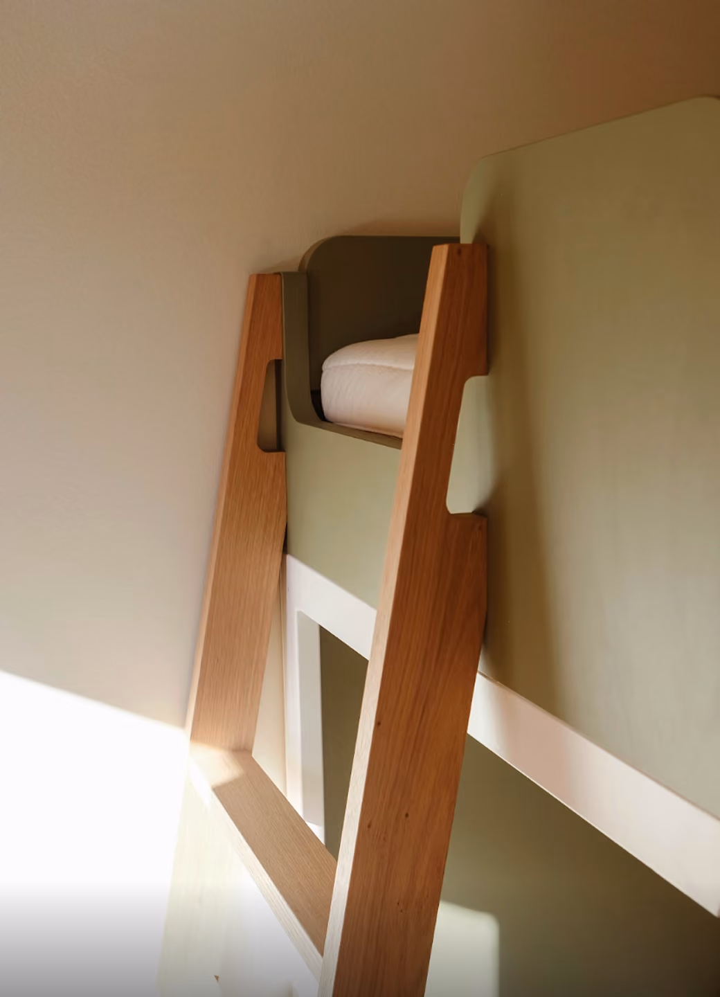 Wooden ladder leading up to a green loft bed with a white mattress in natural light.