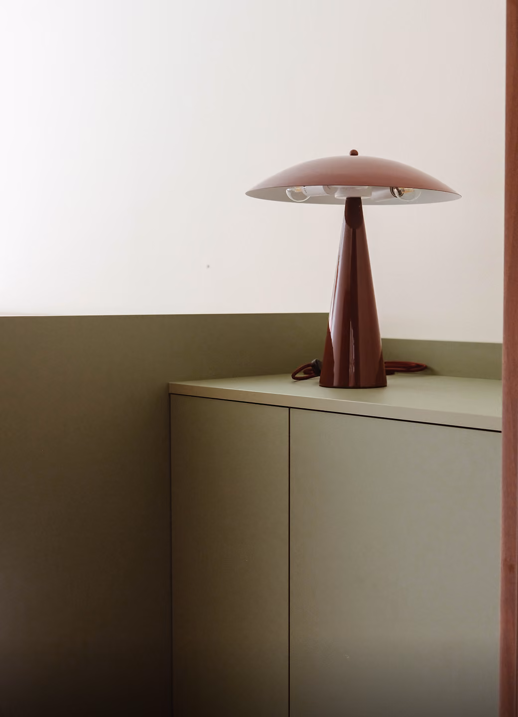Modern brown mushroom-shaped table lamp on a green cabinet against a beige wall.