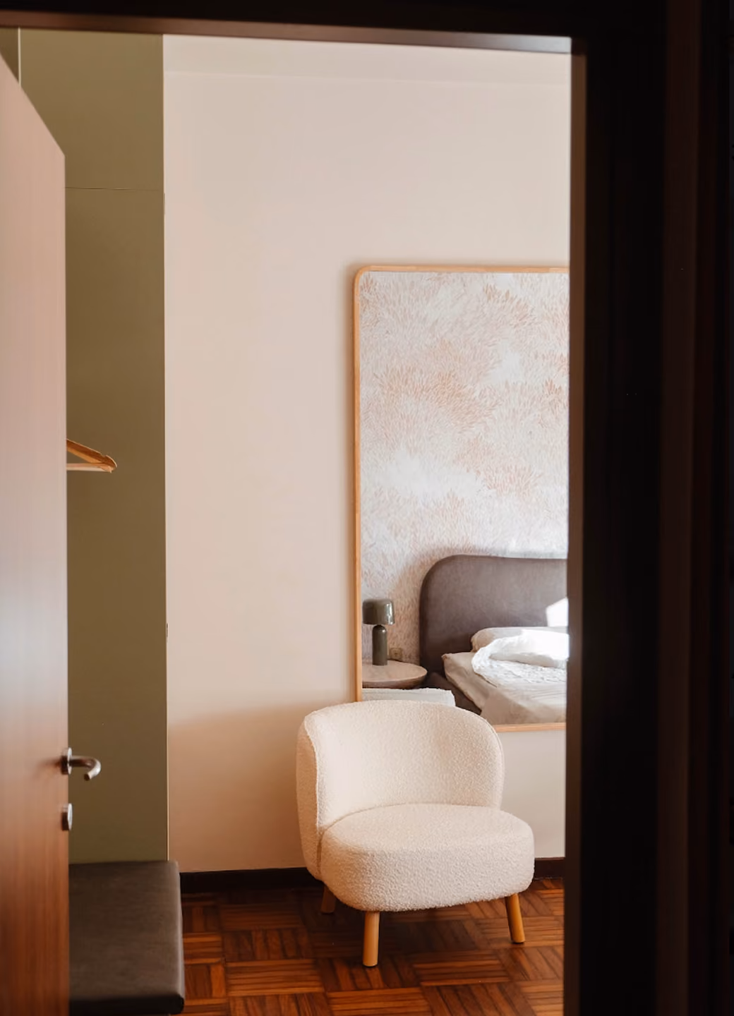 Cozy white armchair on wooden floor in a softly lit bedroom near a large mirror.