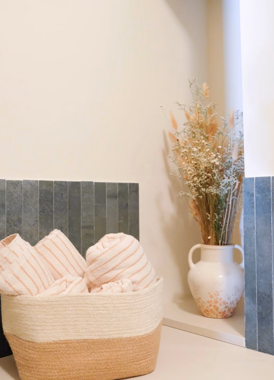 Basket with striped towels next to a white vase of dried flowers on a tiled corner floor.
