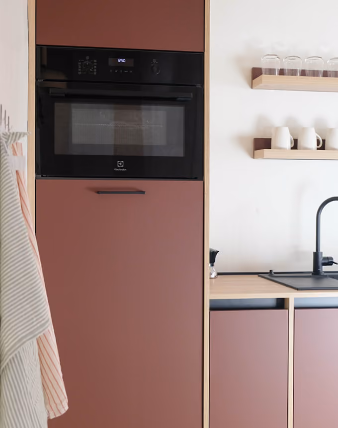 Modern kitchen with built-in black oven and muted red cabinetry beside a black faucet and wooden shelves.