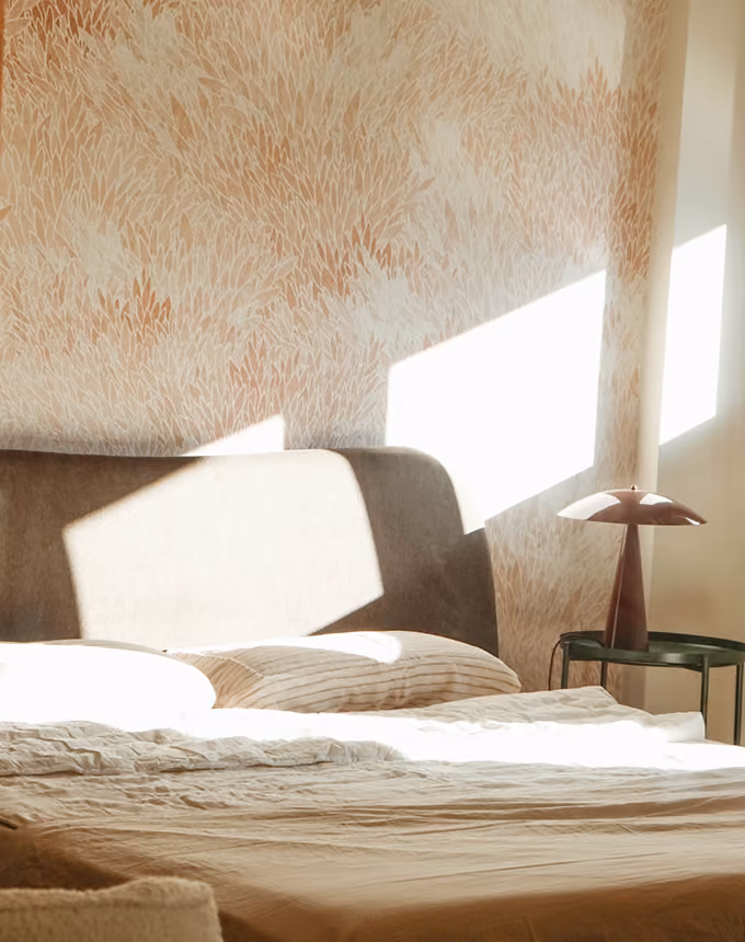 Sunlight casting rectangular shadows on a beige bed with pillows and a bedside table with a lamp.