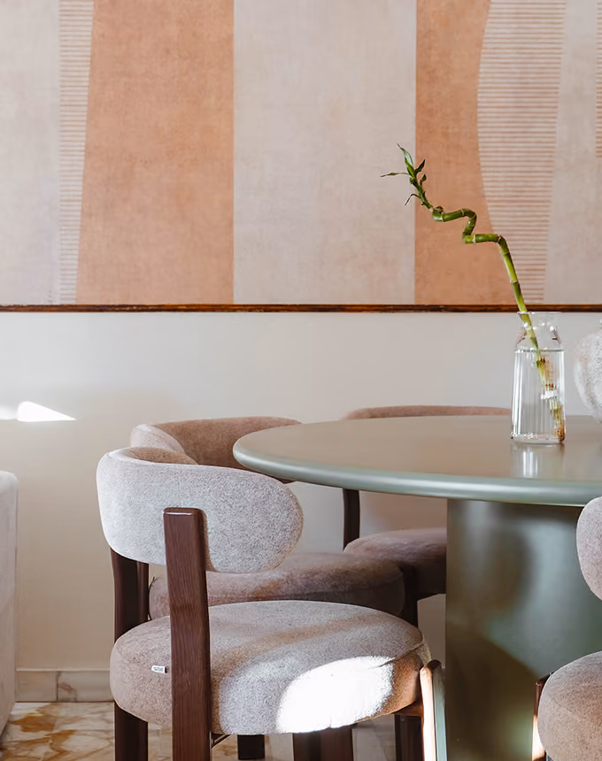 Round table with cushioned chairs and a glass vase holding a curved bamboo shoot in a minimalist room.