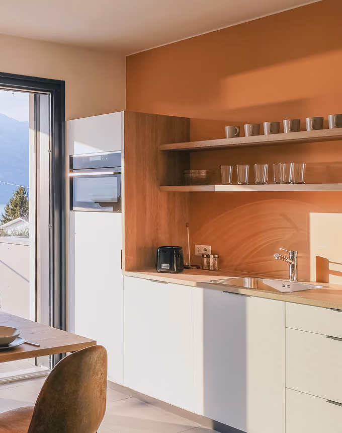 Modern kitchen with white cabinets, wooden shelves holding mugs and glasses, toaster, and sink near a glass door.