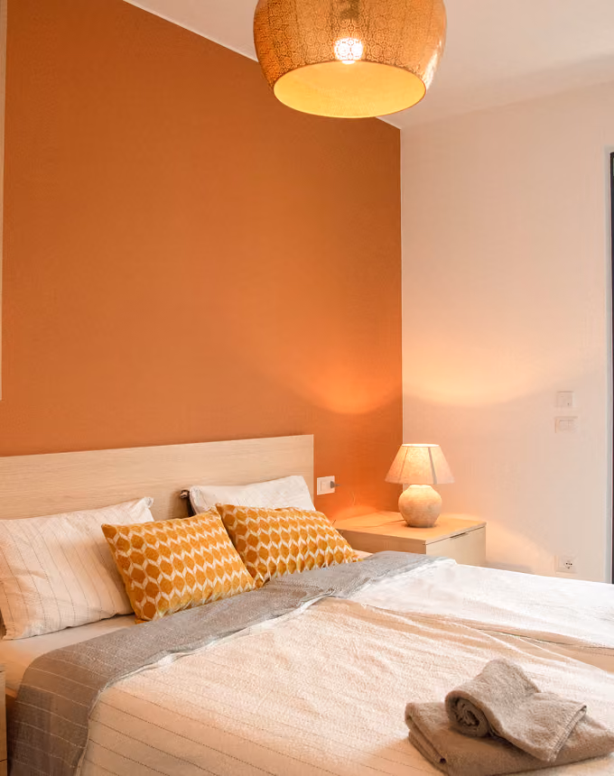 Cozy bed with white and mustard pillows, bedside table with lamp, orange accent wall, and hanging light.