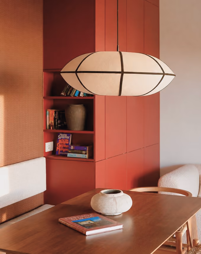 Modern dining area with a wooden table, round vase, unique hanging light, and red built-in shelves with books.