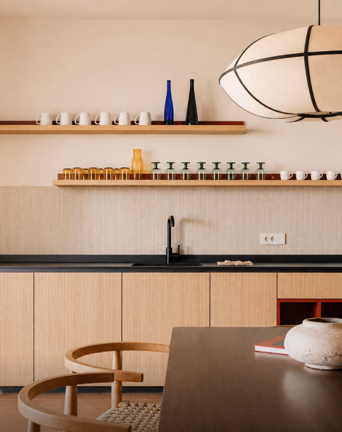 Minimalist kitchen with wooden cabinets, black countertop, floating shelves with cups and bottles, and pendant lamp.