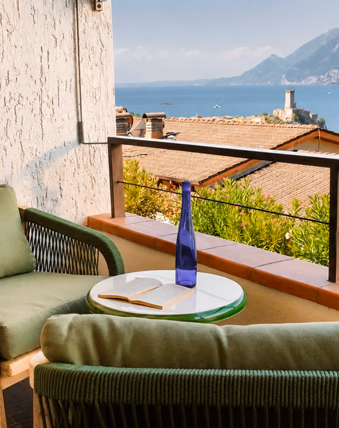 Balcony with green chairs and table with blue bottle and open book overlooking sea and distant castle.