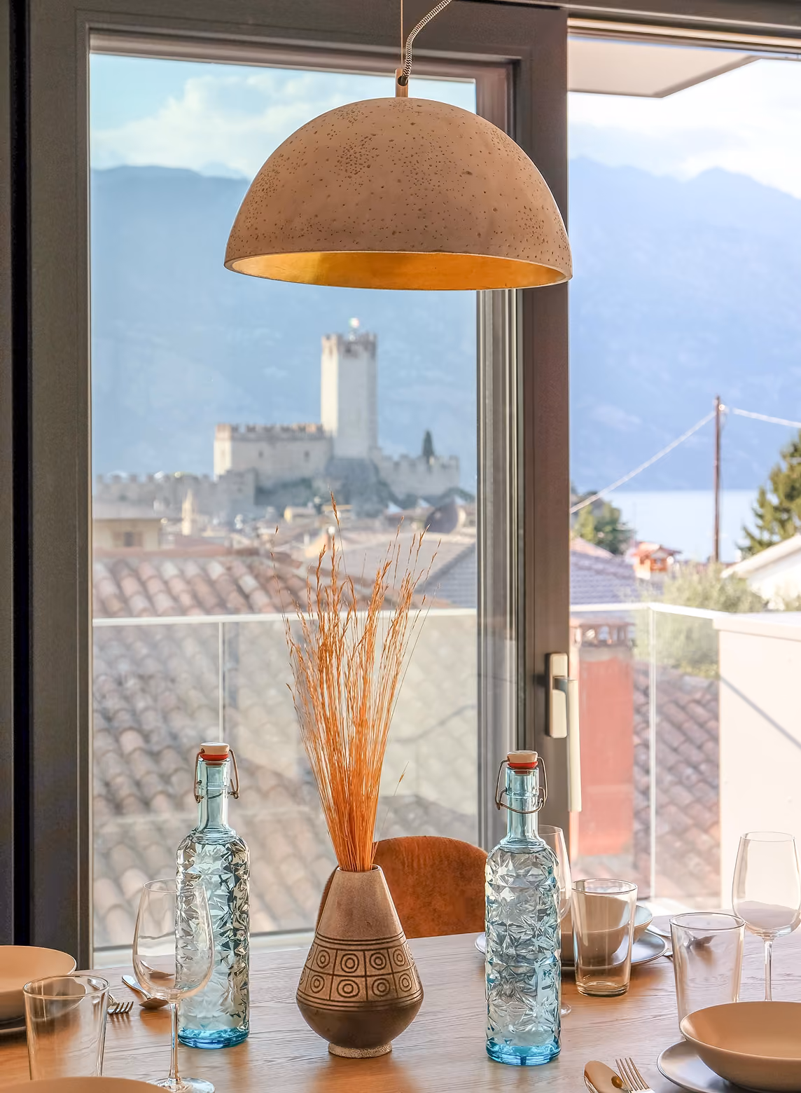 Dining table set with glassware, blue bottles, and vase with dried plants near window showing castle and mountains.