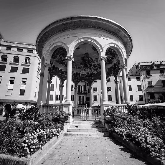 Classical round pavilion with columns in a square surrounded by buildings and flower beds.