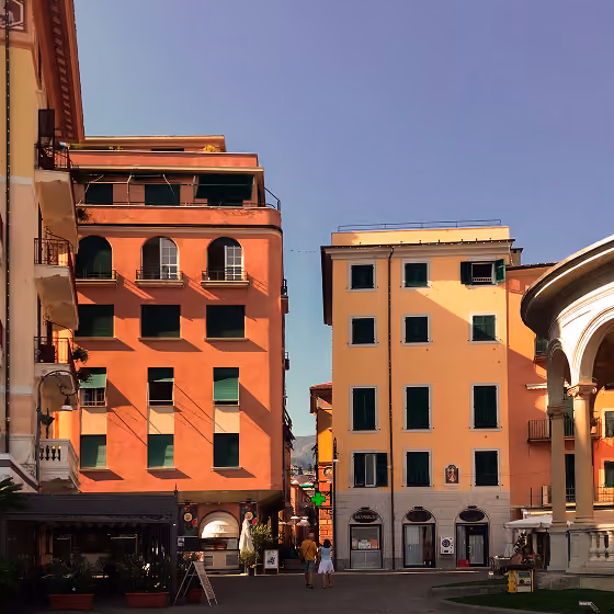 Sunlit orange and yellow buildings with green shutters in a European town square under clear blue sky.
