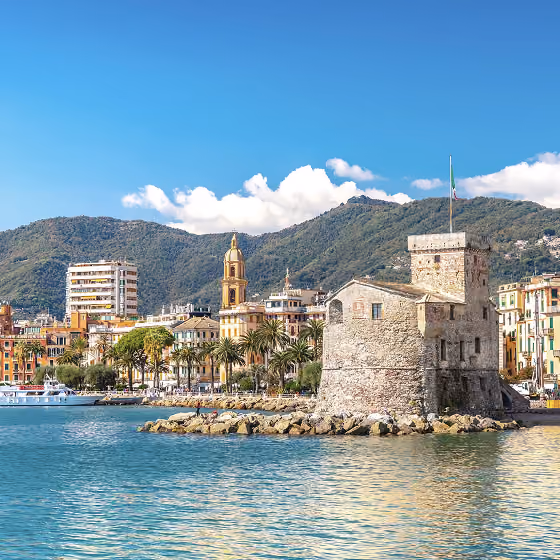 Stone coastal fortress with Italian flag near colorful buildings and mountains under a blue sky.