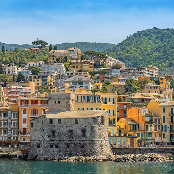 Coastal town with colorful buildings on a hillside and a stone fortress by the water under blue sky.