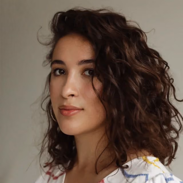 Portrait of a young woman with medium-length curly dark brown hair, wearing hoop earrings and a white top with colorful patterns, looking slightly to the side against a neutral background.