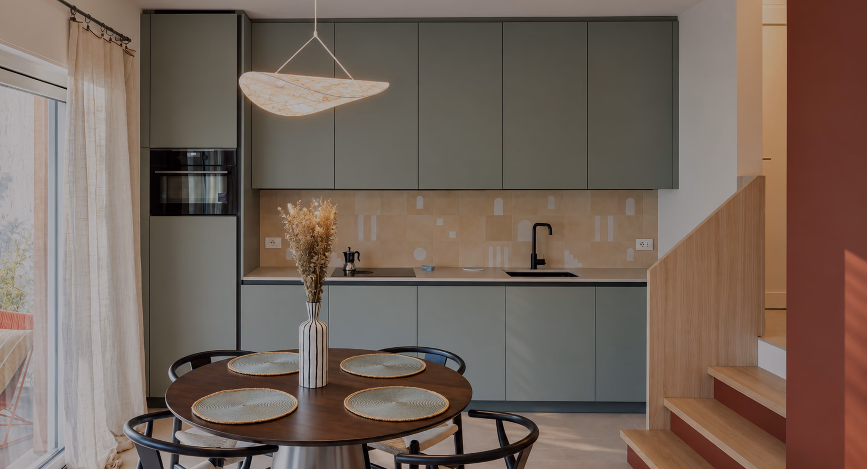 Modern kitchen with matte green cabinets and beige backsplash featuring geometric patterns, a black faucet, and built-in oven. In front, a round wooden dining table set with four woven placemats and a striped vase holding dried plants. Light wooden stairs with a half-wall railing lead up beside the kitchen. Sheer curtains cover a large sliding glass door on the left, letting in natural light.