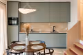 Modern kitchen with green cabinetry, beige backsplash, and countertop holding kitchen appliances, a round dining table with four chairs in front, and wooden stairs adjacent.