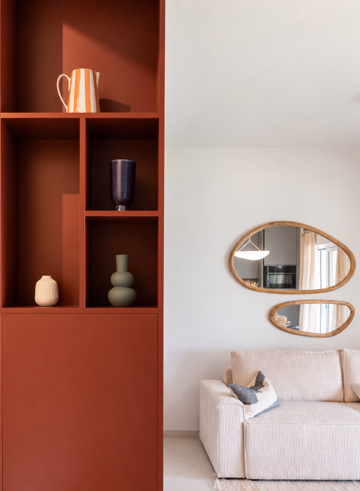 Modern living room corner with a beige ribbed sofa and two decorative pillows next to a white wall featuring two irregular wooden-framed mirrors, and a tall rust-colored shelving unit with four open compartments holding a striped orange and white pitcher, a dark blue vase, a small beige vase, and a matte green vase.