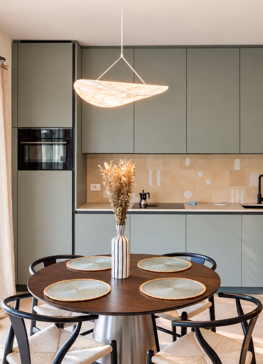 Modern kitchen with muted green cabinets and a round wooden dining table set with four light green placemats and a tall striped vase with dried flowers, illuminated by a uniquely shaped pendant light above.
