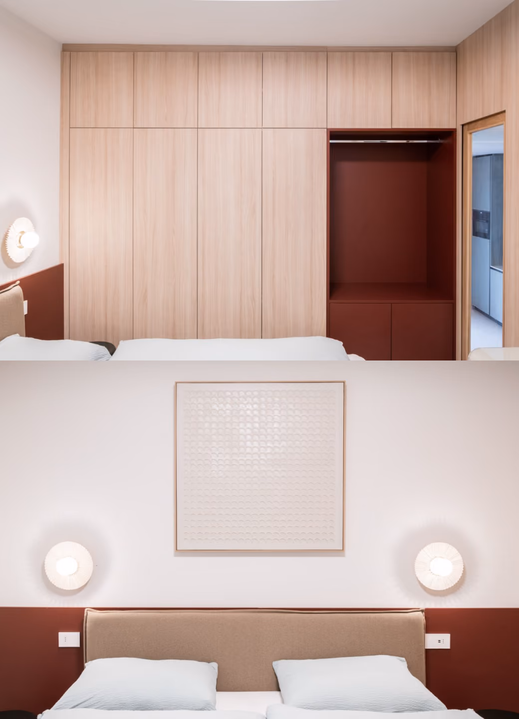 Modern bedroom featuring a large beige upholstered bed with white pillows against a wall half painted dark red. Above the bed is a square textured white artwork framed in light wood, flanked by two round white wall sconces. Opposite the bed is a full wall of light wood cabinets with a dark red open section next to a doorway leading to another room.