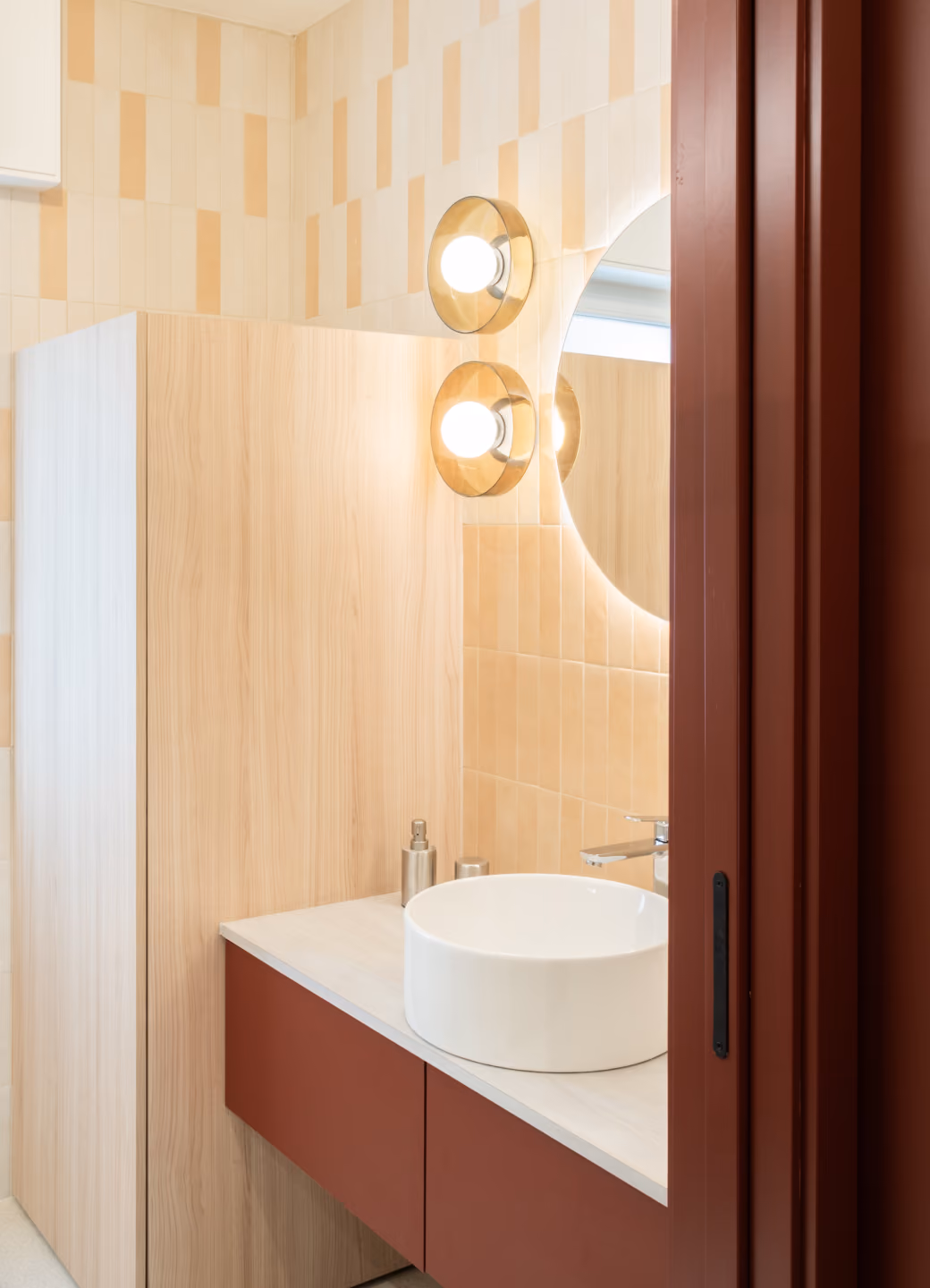 Modern bathroom vanity with a round white vessel sink on a light wood countertop, rust-colored drawers, beige tiled walls, two round wall lights, and a circular mirror reflecting part of a window and the lights.