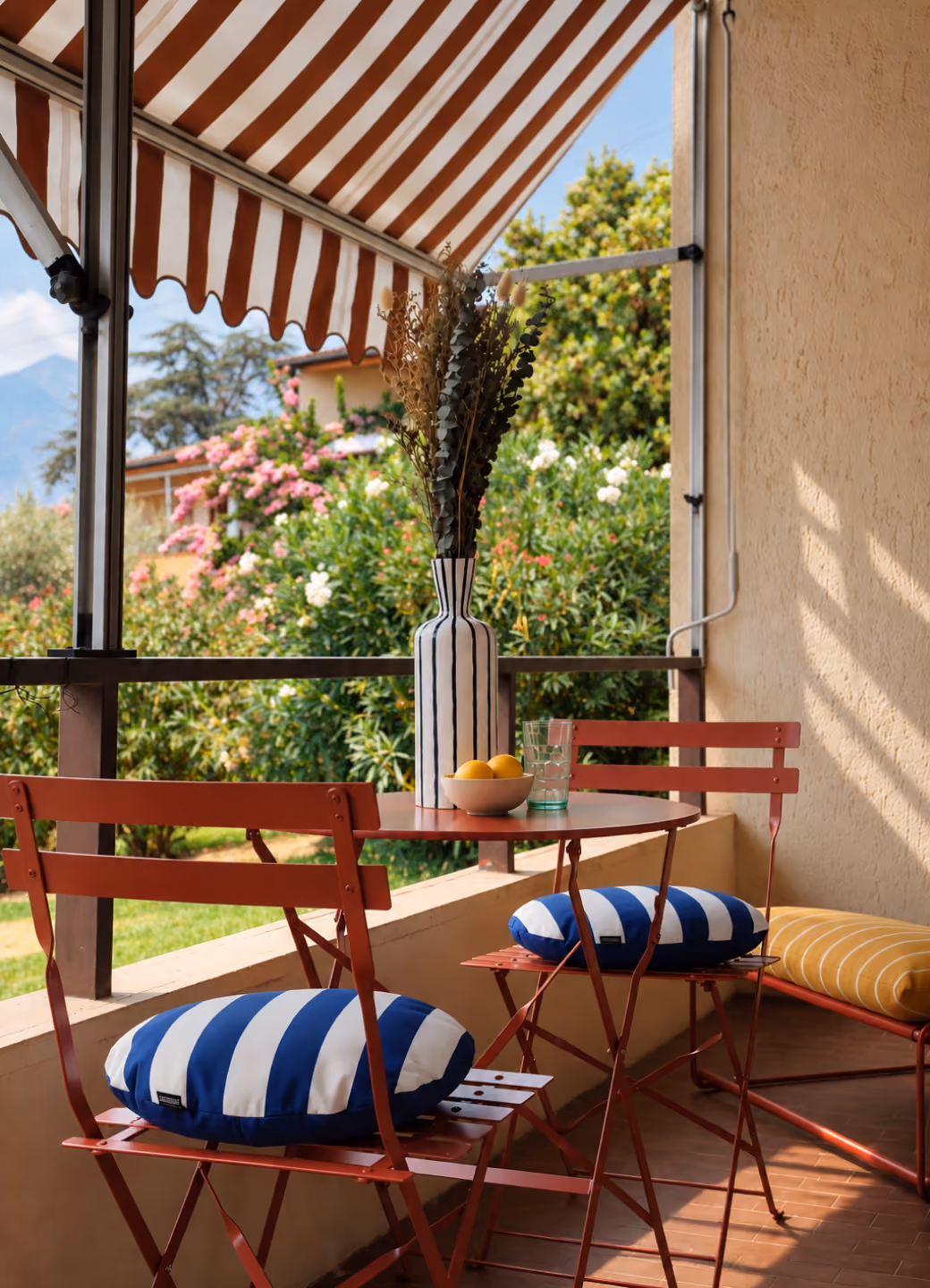 Small outdoor seating area with two red metal chairs with blue and white striped cushions, a red metal round table holding a striped vase with dried plants, a bowl of lemons, and a glass, under a red and white striped awning with green shrubbery and flowers in the background.