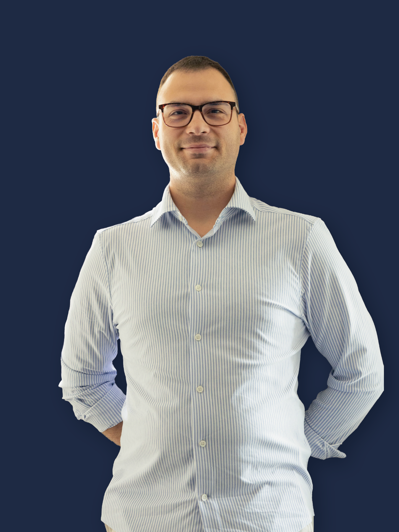 Bărbat cu părul scurt și ochelari, purtând o cămașă cu dungi albastre și albe, cu mâinile la spate pe un fundal albastru închis.