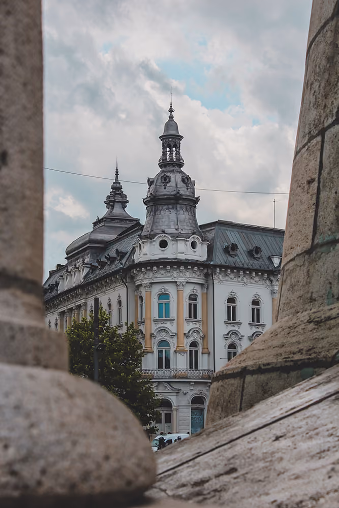 Clădire istorică albă cu ferestre decorative și acoperiș argintiu cu turnuri ornamentale, văzută printr-unghi dintre coloane de piatră, sub cer înnorat.