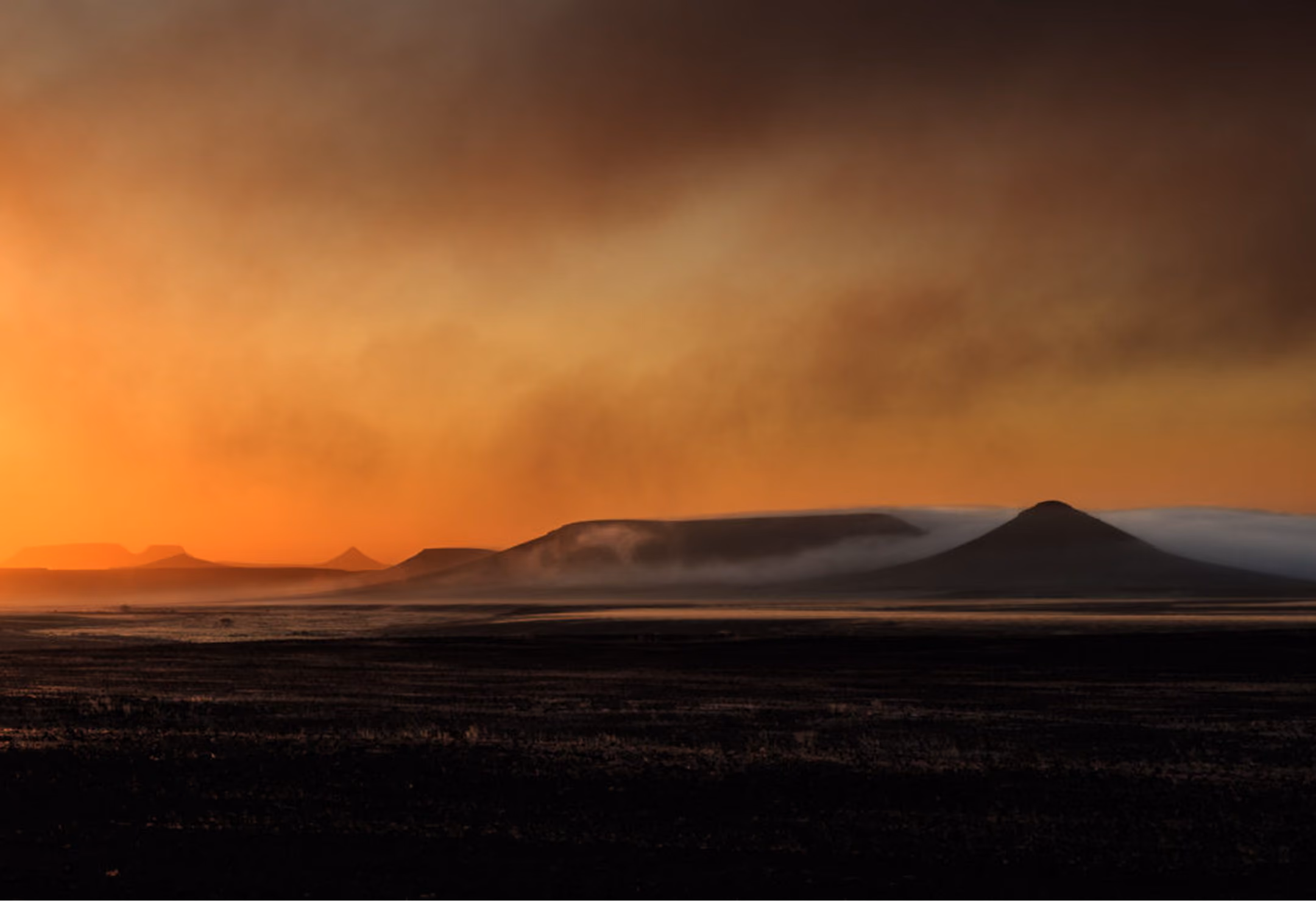 Sunset casting an orange glow over mist-covered hills and flatlands in a dark landscape.