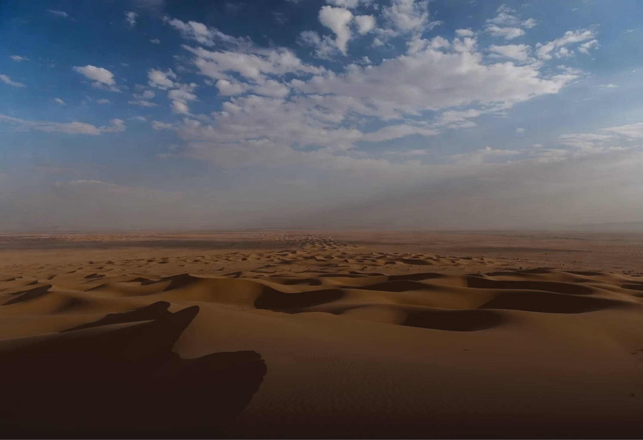 Expansive desert landscape with sand dunes under a partly cloudy blue sky.