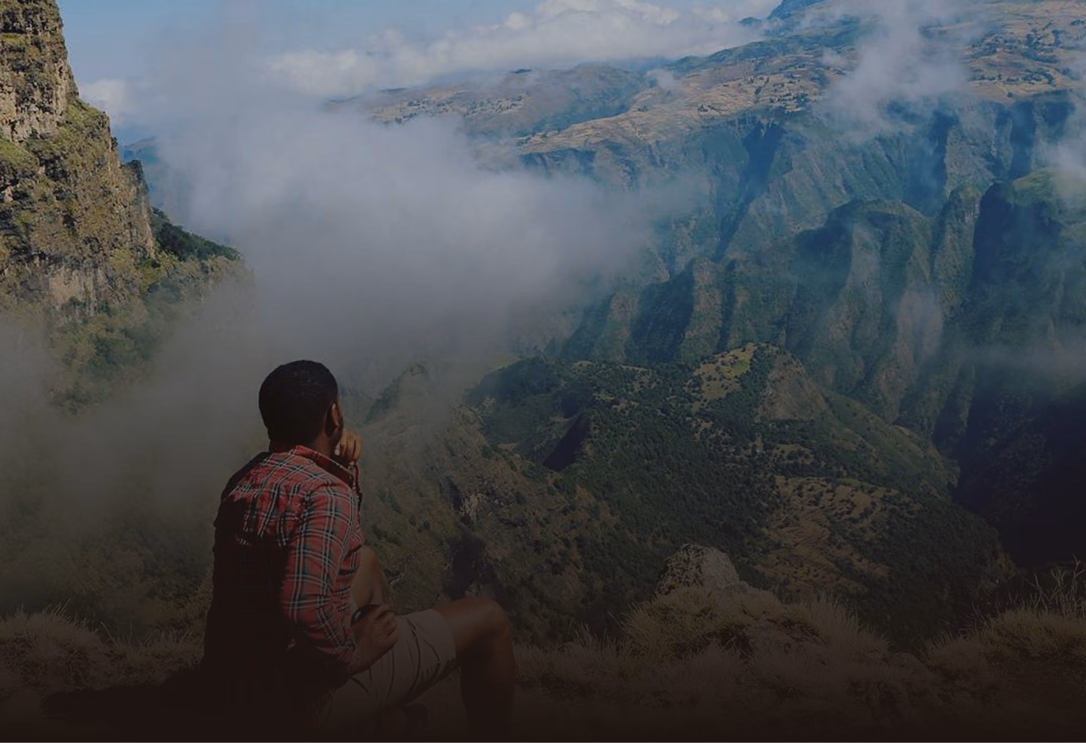 Man in red plaid shirt sitting on a cliff overlooking a foggy mountain valley.